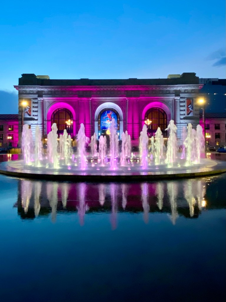 Union Station and the Henry Wollman Bloch Fountain, lit up at night. The colours vary with whatever events are happening within the station. In the photo, the station and water jets in the fountain are mostly pink as the movie 'Barbie' was being shown in the Station's movie theater at the time