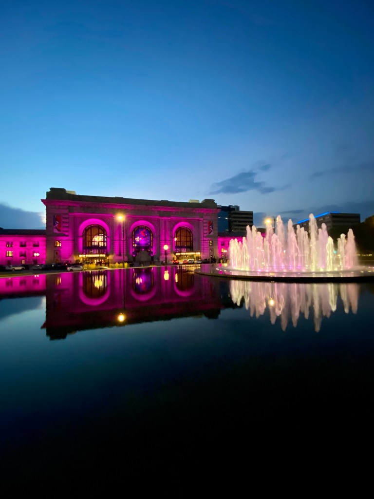 Photo of Union Station and the Henry Wollman Bloch Fountain, lit up at night. When the photo was taken in July 2023, the station and fountain were lit in pink but the colours vary depending on the occasion