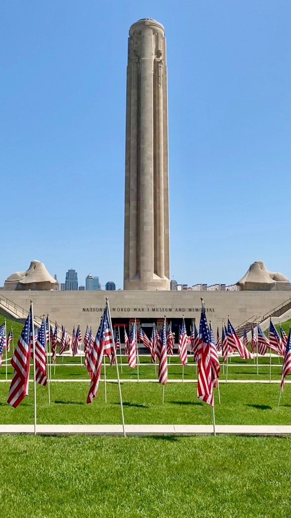 Photo showing the Liberty Memorial tower and, below it, the National World War One Museum in Kansas City. In the foreground are some American flags. In the distance are some of the buildings in downtown Kansas City